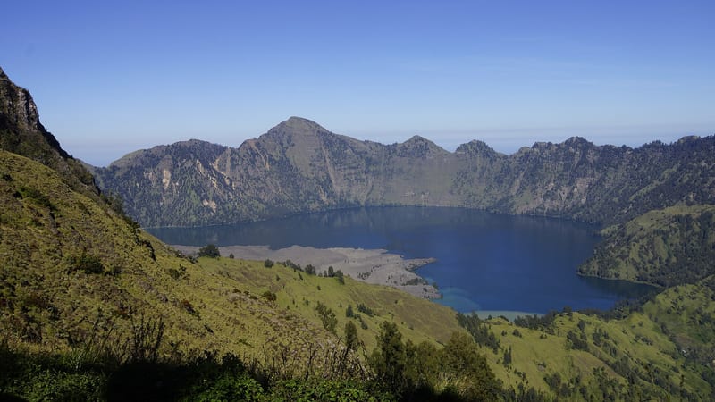 Danau Segara Anak berwarna biru di dalam cekungan Rinjani, dikelilingi tebing dan lereng bervegetasi.