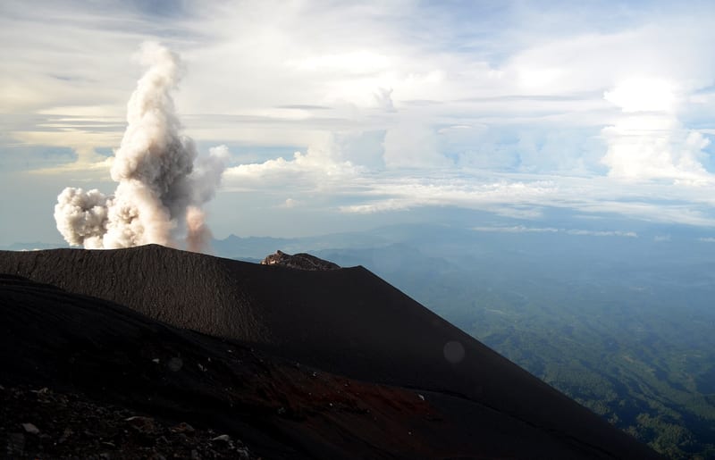 Kepulan material erupsi naik di atas kawah Semeru, dengan lereng gelap dan langit terang.