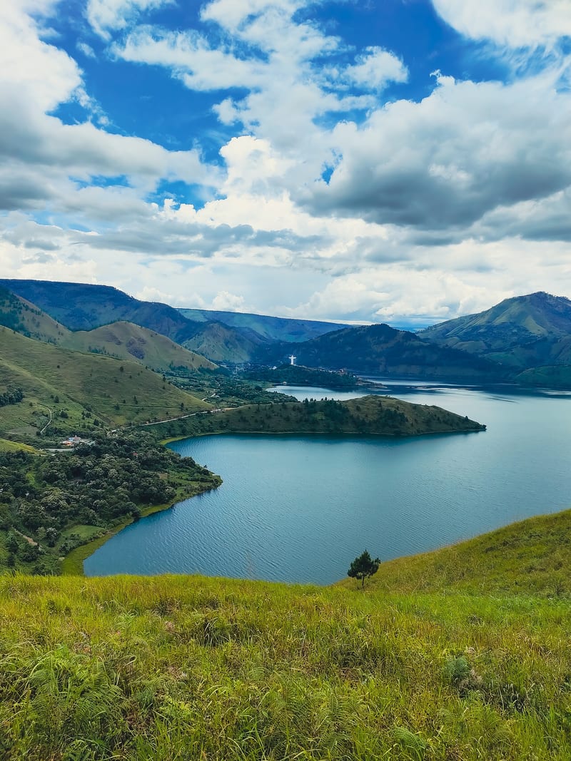 Air Danau Toba diapit lereng hijau dan tanjung, dilihat dari Bukit Holbung di bawah langit berawan.